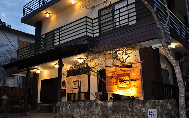 夜の帳が下りる頃、温かな照明に照らされた和モダンな宿「桜ざか」の外観。石垣と木の看板が風情を感じさせる。