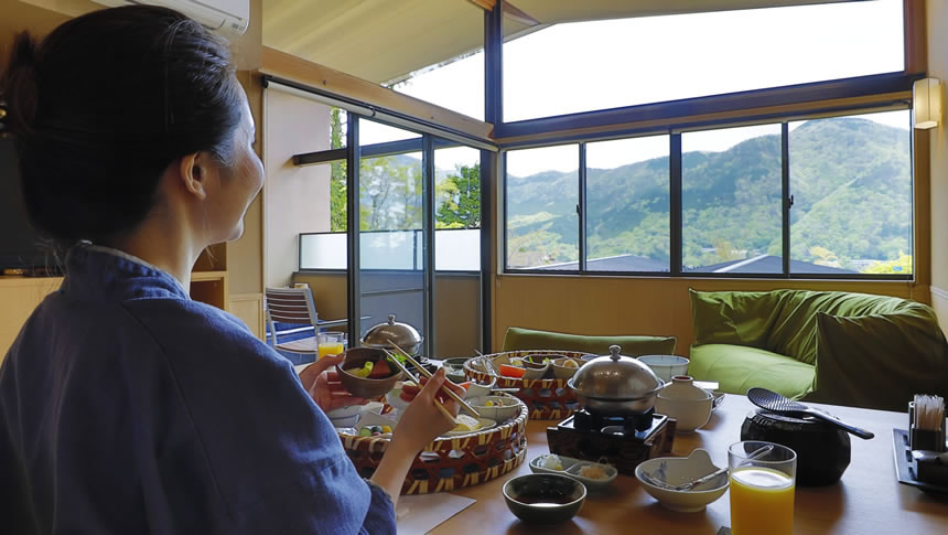 窓いっぱいに広がる山の景色を眺めながら、旅館の部屋で朝食を楽しむ女性