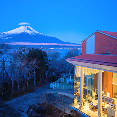 夕暮れ時のレジーナリゾート富士山中湖の外観。ライトアップされた開放的なロビー越しに、冠雪した富士山と山中湖、たなびく笠雲を望む幻想的な風景