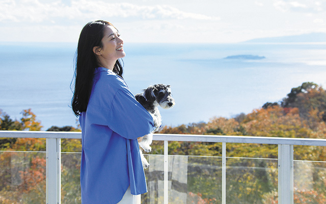 静岡県・熱海の愛犬と泊まれる宿「ゆるり熱海 with DOGS」で愛犬を抱っこして相模湾の絶景を眺める女性。オーシャンビューのバルコニー　。参考価格 18,700円〜